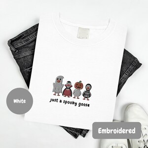 A white t-shirt with a graphic design of three cartoon characters, including a ghost, a pumpkin, and a skeleton, along with the text "Just a spooky goose". The t-shirt is embroidered with the design, and there are jeans and white shoes visible in the background.