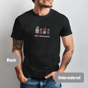 A man wearing a black t-shirt with a graphic of four cartoon characters, including a ghost, pumpkin, skeleton, and a bird, along with the text "just a spooky goose".