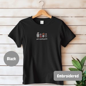 A black t-shirt with a graphic design of four cartoon characters, including a ghost, a pumpkin, and a robot, along with the text "Just a spooky goose" printed on it. The t-shirt is hanging on a wooden hanger against a white background.