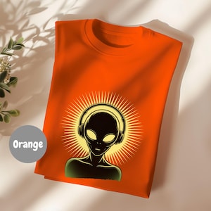 May include: Orange t-shirt featuring a graphic of an alien wearing headphones, set against a radiating sunburst design. The alien has large yellow eyes and a green neckline. The word "Orange" is in a gray circle in the lower left corner.