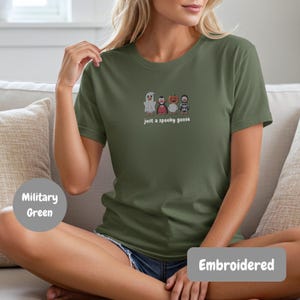 A woman wearing a green t-shirt with a graphic of four cartoon characters, including a ghost, a pumpkin, a skeleton, and a goose, sitting on a couch.