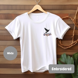 Può includere: Una t-shirt bianca a maniche corte con una piccola grafica sul petto sinistro. La grafica mostra la sagoma nera di un uccello in volo sopra la scritta "Bird Nerd" in nero, con una fila di piccoli fiori colorati sotto il testo.