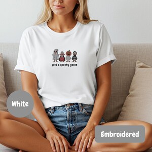 A woman wearing a white t-shirt with a graphic of four cartoon characters and the text "Just a spooky goose" printed on it, sitting on a couch.