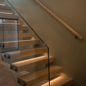 Puede incluir: Escalera de madera moderna con paneles de vidrio y pasamanos de madera. Los escalones están iluminados con iluminación empotrada, creando un ambiente cálido. El diseño es minimalista.