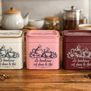 May include: Three square tea tins in cream, pink, and burgundy, each with a floral and teapot design. The text "Le bonheur est dans le thé" is printed on each tin. The tins are on a wooden surface.