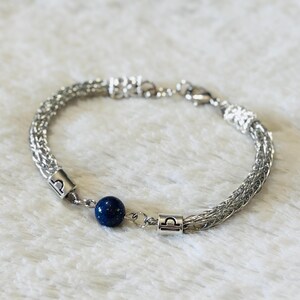 May include: A silver-toned bracelet with a woven chain design. The bracelet features a dark blue, spherical bead and two silver charms with the Libra symbol. The bracelet is displayed on a white, textured surface.
