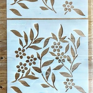 K&ouml;nnte beinhalten: Zwei wei&szlig;e Kunststoff-Schablonen mit einem floralen Design. Die obere Schablone hat eine einzelne Reihe von Blumen und Bl&auml;ttern. Die untere Schablone hat ein aufwendigeres Design mit mehreren Blumen und Bl&auml;ttern.