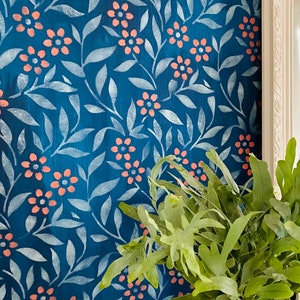 K&ouml;nnte beinhalten: Eine blaue Wand mit einem wei&szlig;en Schablonenmuster aus Bl&auml;ttern und Blumen. Die Blumen sind hellrosa. Eine gr&uuml;ne Pflanze in einem wei&szlig;en Topf befindet sich in der unteren rechten Ecke des Bildes.
