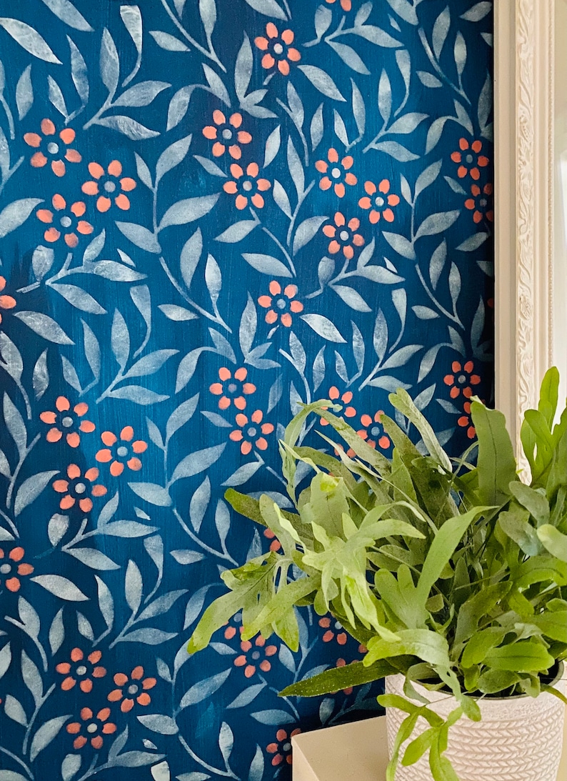 K&ouml;nnte beinhalten: Eine blaue Wand mit einem wei&szlig;en Schablonenmuster aus Bl&auml;ttern und Blumen. Die Blumen sind hellrosa. Eine gr&uuml;ne Pflanze in einem wei&szlig;en Topf befindet sich in der unteren rechten Ecke des Bildes.