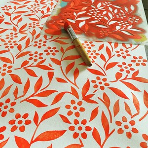 K&ouml;nnte beinhalten: Nahaufnahme eines Schablonen und eines Pinsels, die verwendet werden, um ein sich wiederholendes Muster aus orangefarbenen Bl&auml;ttern und Blumen auf wei&szlig;em Papier zu erstellen.