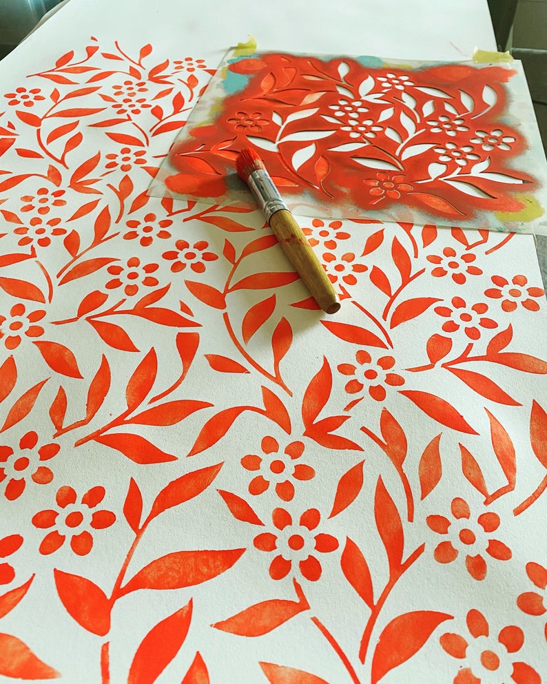 K&ouml;nnte beinhalten: Nahaufnahme eines Schablonen und eines Pinsels, die verwendet werden, um ein sich wiederholendes Muster aus orangefarbenen Bl&auml;ttern und Blumen auf wei&szlig;em Papier zu erstellen.