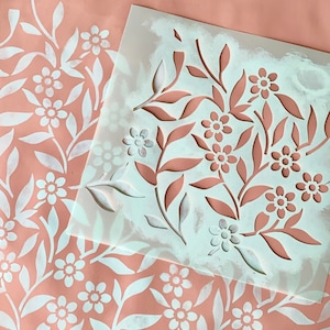 K&ouml;nnte beinhalten: Eine wei&szlig;e Schablone mit einem floralen Muster auf rosa Untergrund. Die Schablone liegt auf einem Blatt Papier, das mit dem gleichen Muster bemalt wurde.