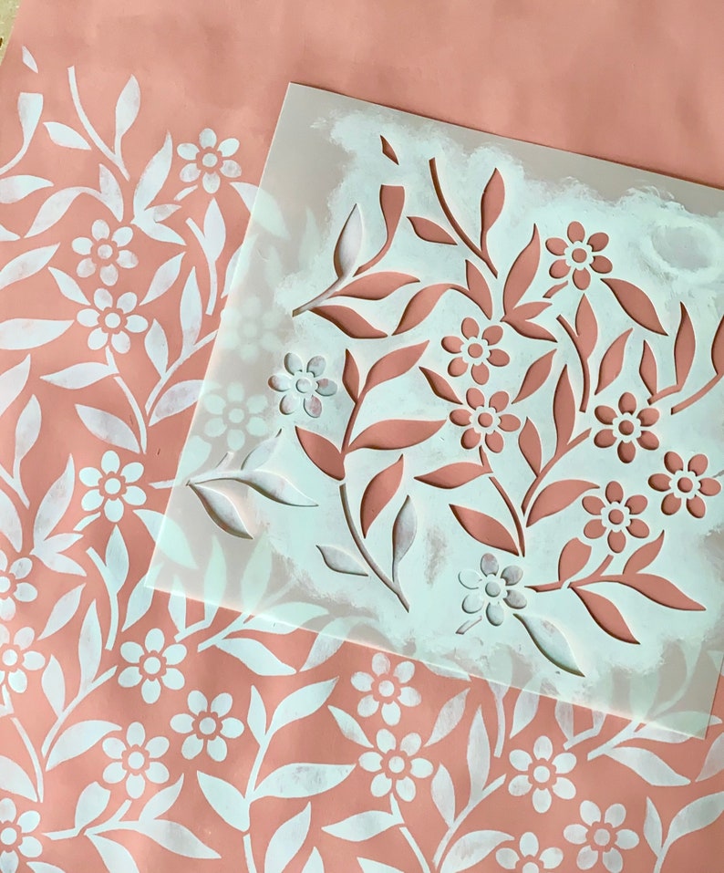 K&ouml;nnte beinhalten: Eine wei&szlig;e Schablone mit einem floralen Muster auf rosa Untergrund. Die Schablone liegt auf einem Blatt Papier, das mit dem gleichen Muster bemalt wurde.