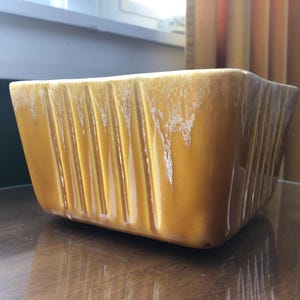 May include: A yellow ceramic planter with vertical lines and a white glaze. The planter is sitting on a wooden surface.
