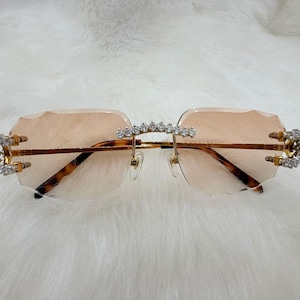 May include: Rimless sunglasses with peach-tinted lenses and a gold-colored frame. The temples and bridge are adorned with sparkling rhinestones. The arms have a tortoise shell pattern.