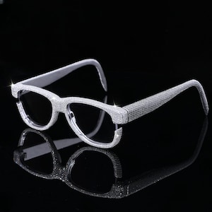 May include: Silver-toned eyeglasses encrusted with sparkling rhinestones. The frames and temples are fully covered in the shimmering embellishments, reflecting light. The lenses are clear, and the glasses are set against a dark, reflective surface.