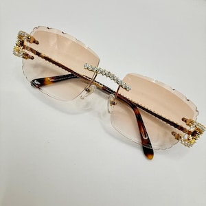 May include: Rimless sunglasses with a light pink tint. The frames are gold-colored with decorative rhinestones along the top and at the temples. The arms are a tortoiseshell pattern.