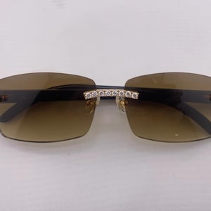 Könnte beinhalten: Rahmenlose Sonnenbrille mit braunen Verlaufsgläsern. Die Fassungen sind schwarz mit goldenen Akzenten und mit kleinen, klaren Kristallen besetzt. Der Steg und die Bügel sind ebenfalls mit Kristallen verziert.