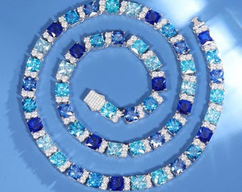 Könnte beinhalten: Eine schillernde Halskette mit einem spiralförmigen Design aus quadratisch geschliffenen blauen und klaren Edelsteinen. Die Halskette zeigt einen Farbverlauf von Blautönen, von helltürkis bis tiefnavy, in einem silberfarbenen Metall. Der Verschluss ist ebenfalls mit klaren Steinen besetzt.