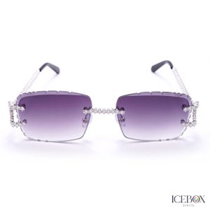 Può includere: Occhiali da sole rettangolari con lenti sfumate viola. Le montature e le aste sono argentate e incastonate con piccoli strass trasparenti. Design moderno e lussuoso.