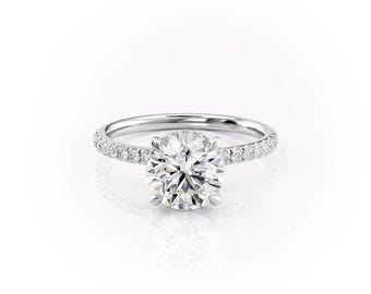 Könnte beinhalten: Ein funkelnder Verlobungsring mit einem großen, rund geschliffenen Diamanten als Mittelpunkt. Das Band besteht aus weißem Metall und ist mit kleineren Diamanten besetzt. Der Ring ist vor weißem Hintergrund platziert.