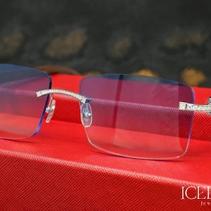 Könnte beinhalten: Randlose Brille mit blau getönten Gläsern und Diamantbesatz am Rahmen. Die Bügel sind aus dunklem Holz. Die Brille wird auf einer roten, strukturierten Oberfläche präsentiert. Der Text "ICEBOX Jewels" ist sichtbar.