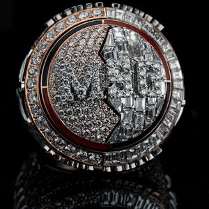 Puede incluir: Un anillo grande y ornamentado con un diseño circular. El anillo está incrustado con numerosos diamantes y presenta un diseño central con las letras "MLB" en una fuente estilizada. El anillo tiene una combinación de colores de dos tonos con detalles en plata y oro rosa.