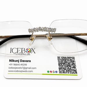 Può includere: Occhiali senza montatura con struttura in metallo dorato e lenti trasparenti. Il ponte e le cerniere sono decorati con file di pietre bianche scintillanti. Sotto è posizionato un biglietto da visita con il testo ICEBOX JEWELS PVT. LTD., Nikunj Davara, contatti e un codice QR.