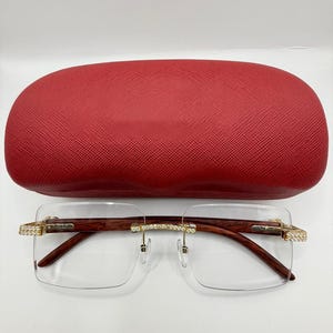 May include: Rimless eyeglasses with brown wooden arms and gold accents, including rhinestones at the temples and bridge. The glasses are paired with a red, textured eyeglasses case.