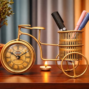 May include: A gold-colored metal bicycle-shaped desk organizer with a clock face and pen holder. The clock has Roman numerals and the Harley-Davidson logo. Pens and a small candle are visible.