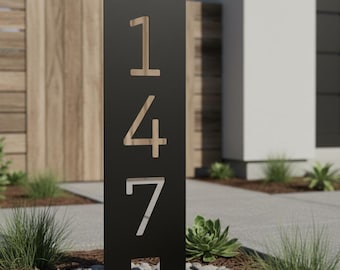 Puede incluir: Un moderno letrero de dirección negro con el número 147 recortado. Los números son de color madera clara, y el letrero está montado en un jardín con pequeñas rocas y vegetación. El letrero es un diseño contemporáneo para una casa.