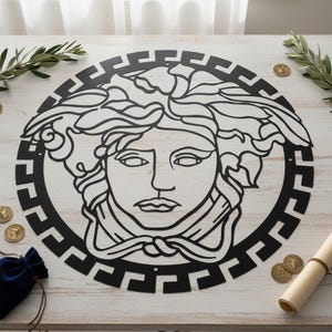 May include: Black metal wall art featuring a Medusa head design within a circular frame. The artwork is placed on a white wooden surface, with decorative elements like coins and greenery surrounding it. The design is a bold, graphic statement.