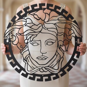 May include: Black metal wall art featuring a stylized Medusa head within a circular frame. The design includes intricate details of the face, hair, and a geometric border. The artwork is held up in front of a light-colored background.
