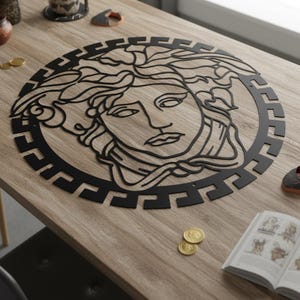 May include: Black metal wall art featuring a stylized Medusa head within a circular frame. The design is intricate, with detailed lines and a geometric border. The artwork is displayed on a light wood surface.