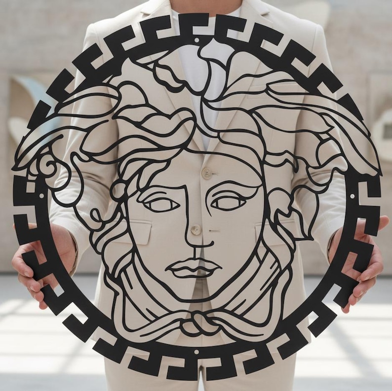 May include: A black metal wall art piece featuring a Medusa head design within a circular frame. The artwork has intricate details and a Greek key border. The piece is held up in front of a neutral background.