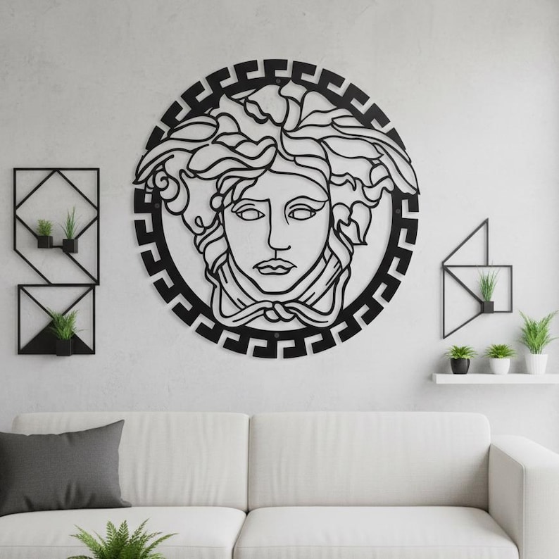 May include: Black metal wall art featuring a Medusa head design within a circular frame. The artwork is mounted on a white wall, with geometric plant holders and a white sofa in the foreground. The Medusa head is a detailed, stylized depiction.