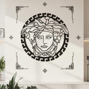 May include: Black metal wall art featuring a stylized Medusa head within a circular frame. The design includes a Greek key border and decorative corner elements, creating a bold, graphic statement. The artwork is mounted on a white wall.