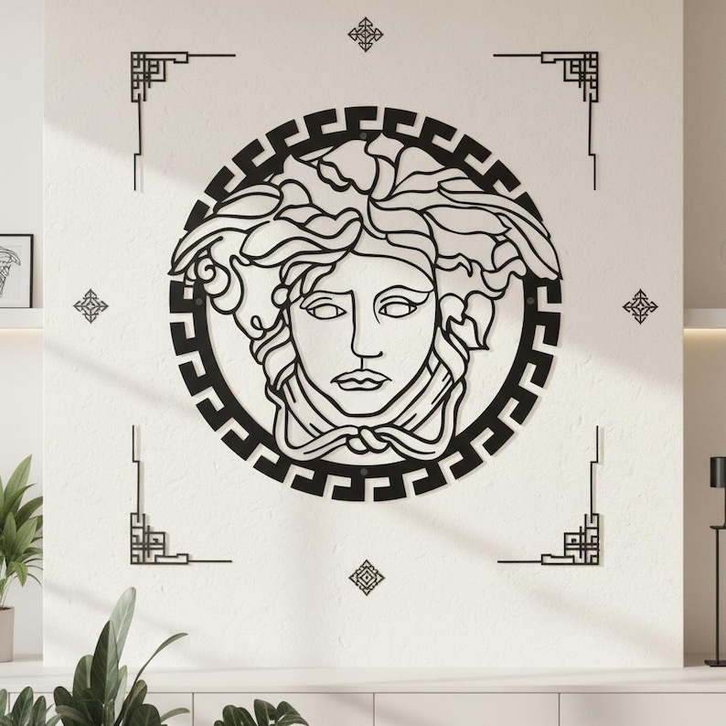 May include: Black metal wall art featuring a stylized Medusa head within a circular frame. The design includes a Greek key border and decorative corner elements, creating a bold, graphic statement. The artwork is mounted on a white wall.
