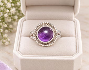 May include: A silver ring featuring a round, polished purple amethyst cabochon stone set in the center. The stone is surrounded by a decorative border of twisted rope and small silver beads, with a split shank band design.