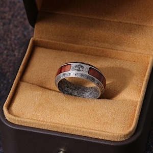 May include: A silver ring with a dark wood inlay and engraved detailing, presented in a brown jewelry box with a tan interior. The ring features a tree symbol and runes.