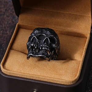 May include: A dark, skull-shaped ring with intricate details, displayed in a brown jewelry box with a tan interior. The ring is made of a dark metal and has a detailed skull design. The box is closed and has a gold-colored button.