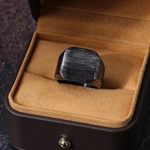 May include: A dark grey, square-faced ring with a brushed metal texture sits in a brown jewellery box with a tan interior. The ring has a flat top and rounded edges. The box is open, displaying the ring.