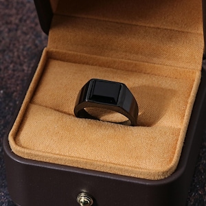 May include: A black ring with a rectangular black stone set in a black band, displayed in a brown ring box with a tan interior. The ring has a modern, minimalist design.