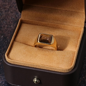 May include: A gold ring with a rectangular, brown gemstone set in a gold bezel. The ring is displayed inside a brown jewelry box with a tan interior. The box is open, showcasing the ring.