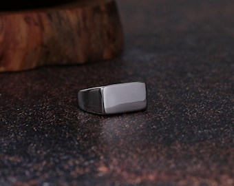 May include: A silver-colored rectangular ring. The ring has a smooth, polished surface and a simple, modern design. The ring is set against a dark, textured background.