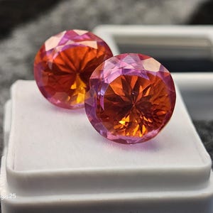 Op de afbeelding: Twee ronde, gefacetteerde edelstenen met een kleurengradiënt van oranje, roze en geel. De edelstenen liggen op een wit oppervlak, met een vage achtergrond. Geschikt voor het maken van sieraden.