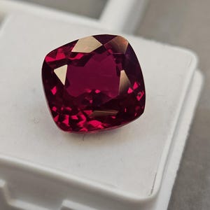 Op de afbeelding: Een dieprode edelsteen met kussenslijpsel en facetten die het licht weerkaatsen. De edelsteen ligt op een wit oppervlak en toont zijn levendige kleur en helderheid. Voor sieraden.