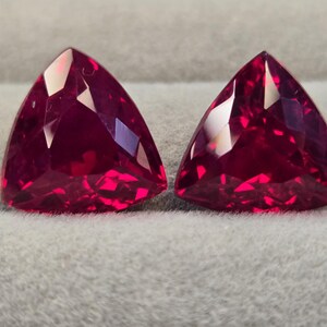 May include: Two triangular-cut, faceted gemstones with a deep red hue. The gems have a polished surface, reflecting light and creating a brilliant sparkle. The gems are set against a neutral grey background.