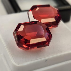 May include: Two faceted, reddish-orange gemstones with an octagonal shape are displayed on a white surface. The gems are highly reflective, catching the light and showcasing their internal structure. The background includes a black box.