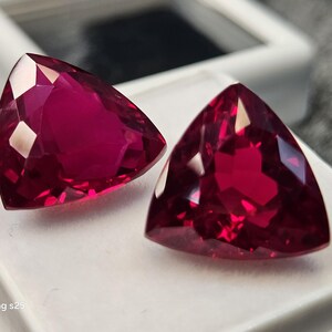 May include: Two triangular, faceted, deep red gemstones. The gems are displayed on a white surface, with a blurred background. The gems have a high luster and reflect light, showcasing their internal structure. The image is well-lit, highlighting the colour and clarity of the stones.